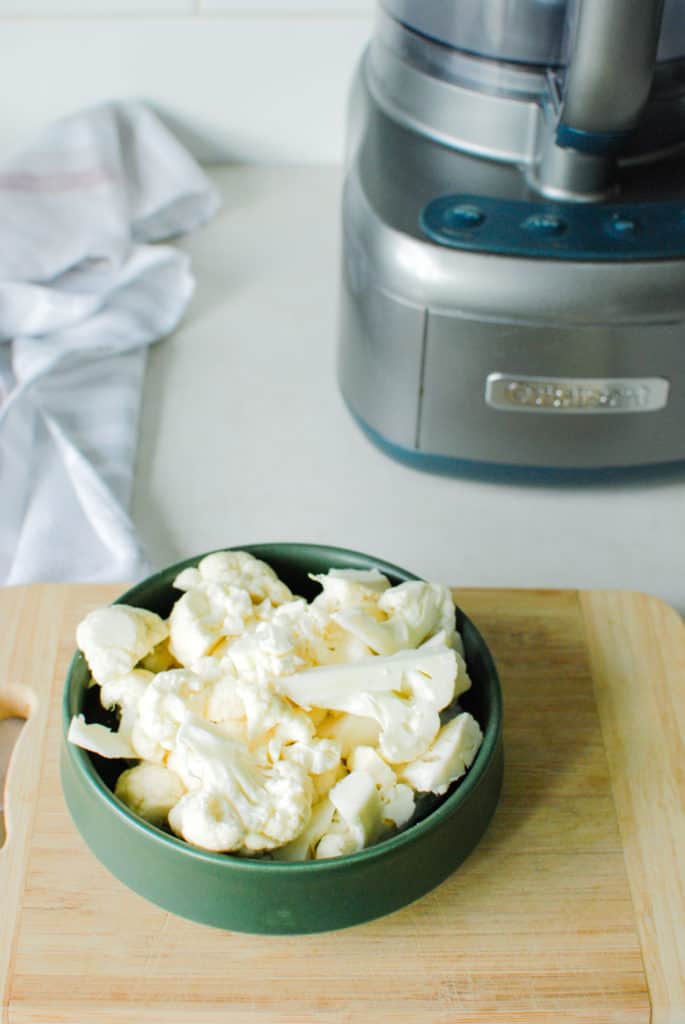 cauliflower florets chopped on a cutting board near a food processor