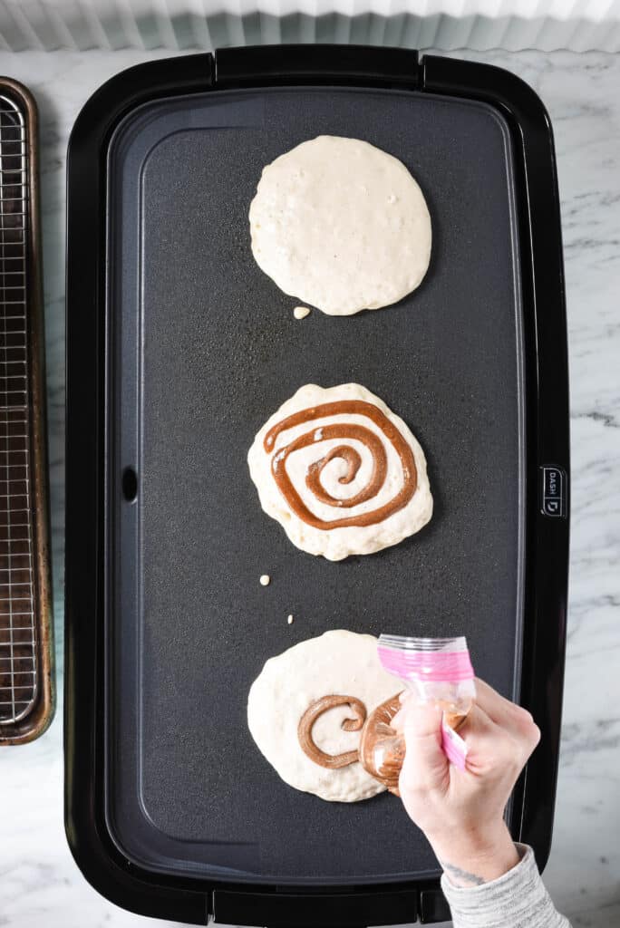 A griddle with 3 cinnamon roll pancakes cooking and a hand pouring a swirl onto each pancake.