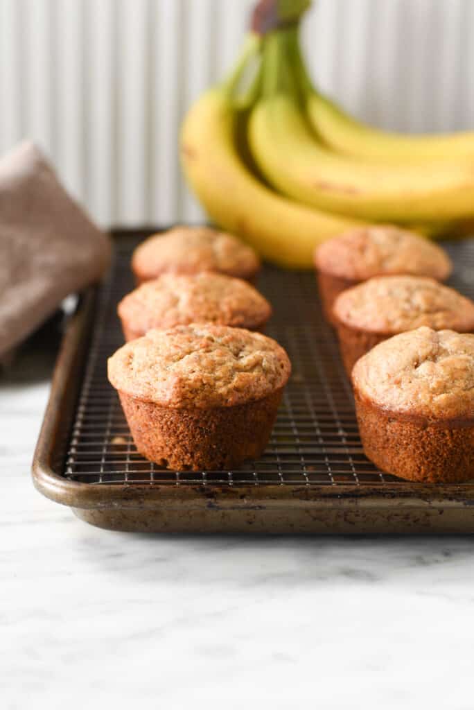 Plant based banana muffins on a cooling rack next to a bundle of fresh bananas.