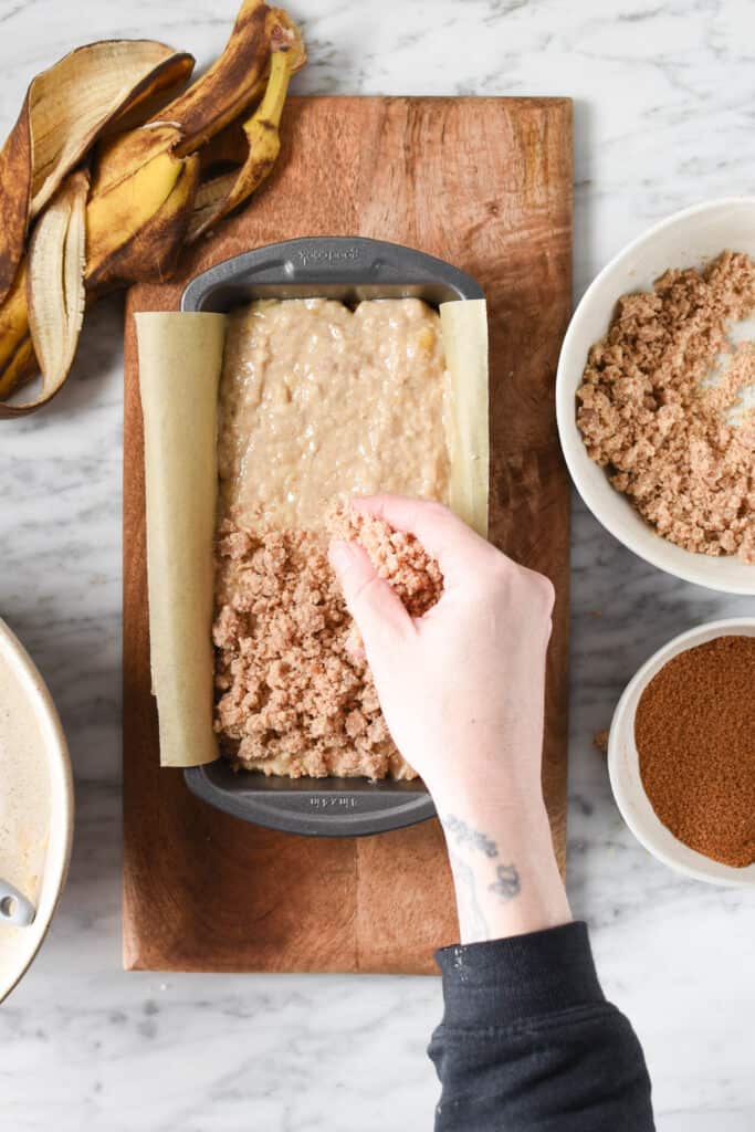 A hand adding a streusel crumb topping to a pan of banana bread batter.